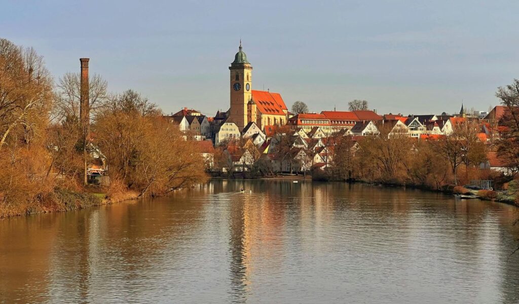 A picturesque riverside town with historic buildings, a tall clock tower, and calm waters reflecting the charming scenery under a clear sky.