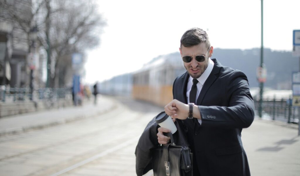 Man in a suit and sunglasses checking his watch while standing near a train track, holding a briefcase and coffee cup.