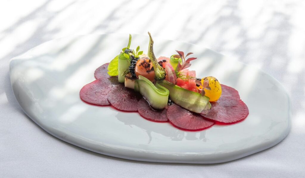 A beautifully plated gourmet dish featuring thinly sliced beets, fresh vegetables, microgreens, and artistic garnishes on a white ceramic plate.