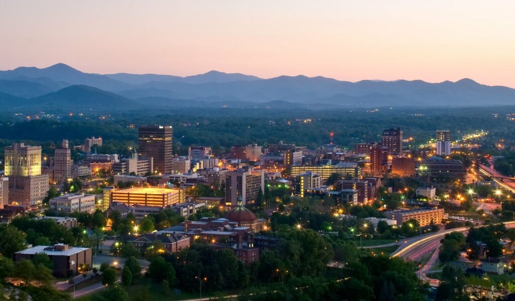 Evening view of downtown Asheville, North Carolina, with city lights glowing beneath layers of Blue Ridge Mountains.