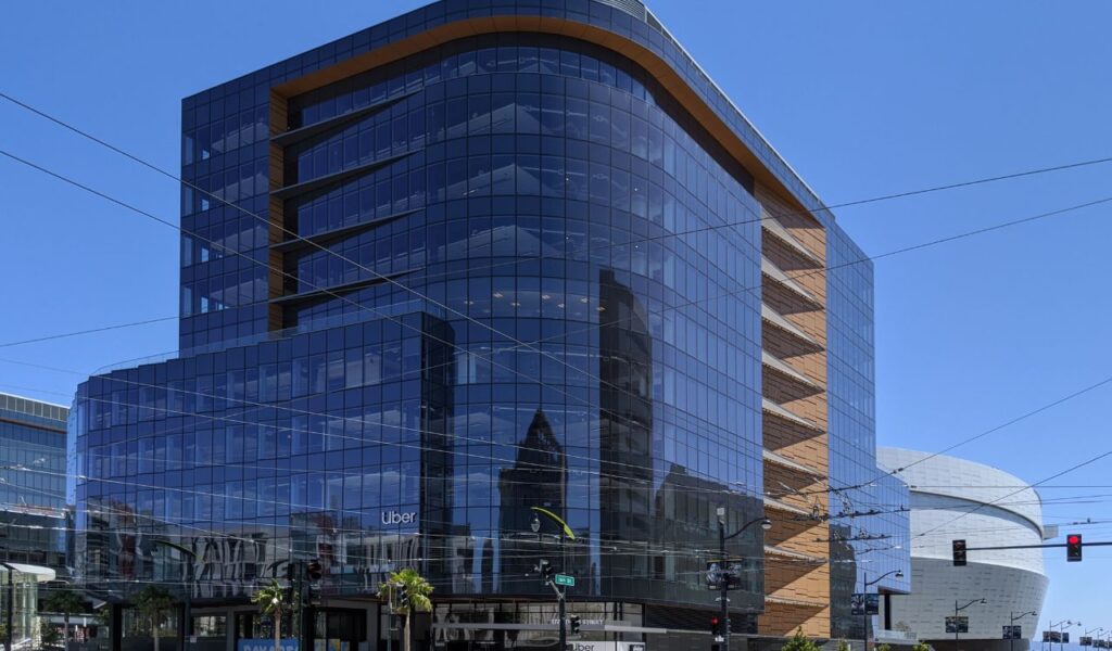 The image shows a modern Uber office building with large glass windows, featuring a sleek design and the Uber logo. A futuristic, curved building is visible in the background.