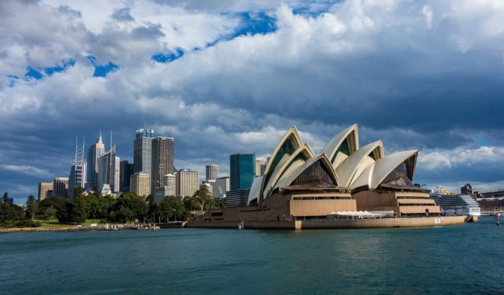 Iconic view of the Sydney Opera House with its sail-like design, set against the modern skyline of Sydney, Australia, under dramatic cloudy skies.