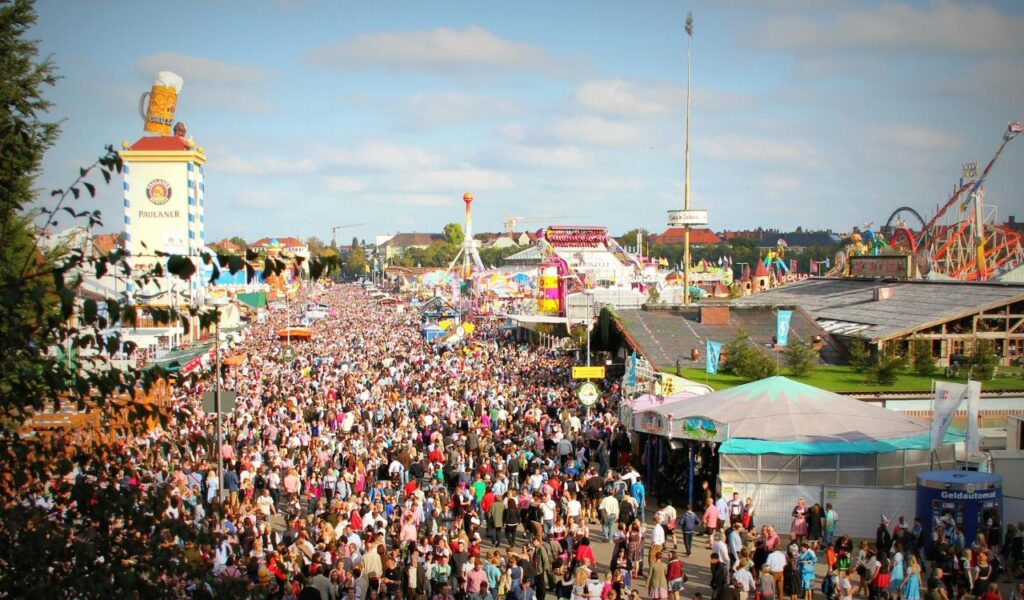 Large crowd at Oktoberfest in Munich, Germany, with colorful tents, traditional attire, and amusement rides visible.
