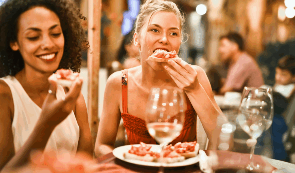 Two women enjoying bruschetta and wine at a cozy, dimly lit restaurant, suggesting a relaxed and authentic local dining experience away from tourist crowds.