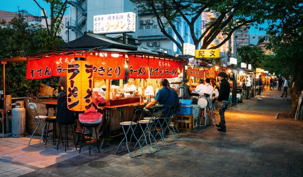 A bustling row of open-air ramen stalls glows under red lanterns in a Japanese city at dusk, with locals enjoying meals and conversations.