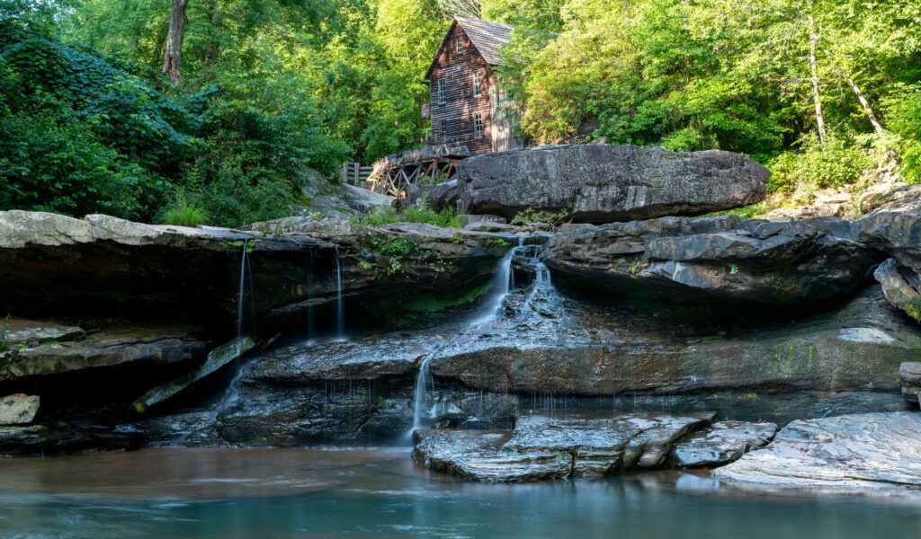 A historic wooden grist mill stands above layered rocks and a small cascading waterfall surrounded by lush green forest in Babcock State Park, Clifftop.