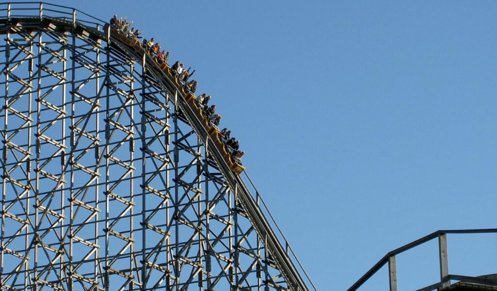 People riding a wooden roller coaster at Bakken Amusement Park in Denmark, with the coaster descending a steep track under a clear blue sky.
