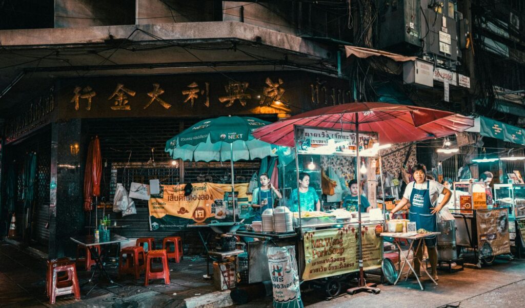 Night street food market in Bangkok with vendors preparing dishes under colorful umbrellas and illuminated stalls.