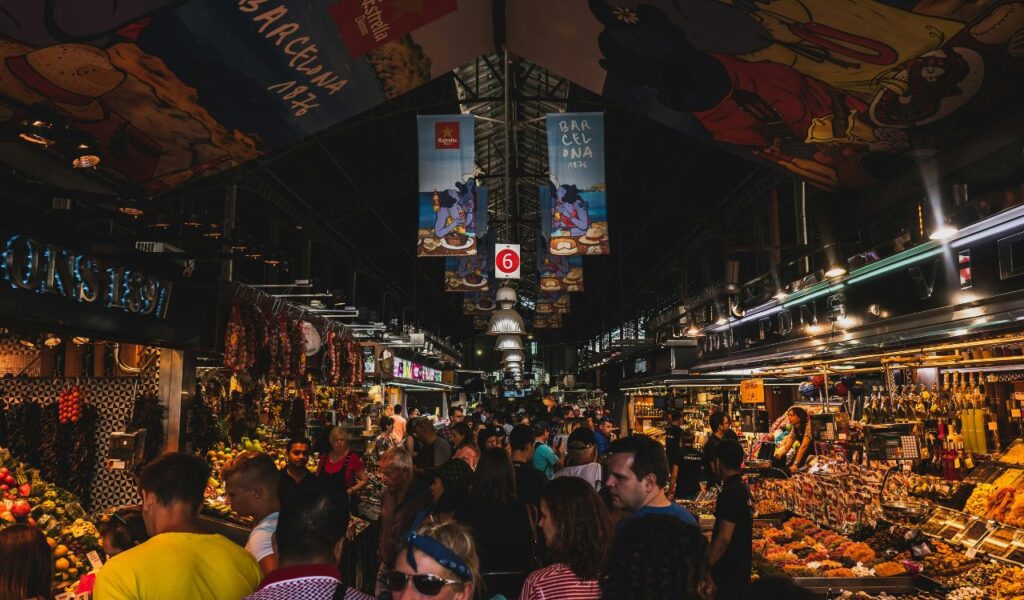 Crowded indoor food market in Barcelona with stalls displaying fruits, meats, and local products under colorful banners and lights.