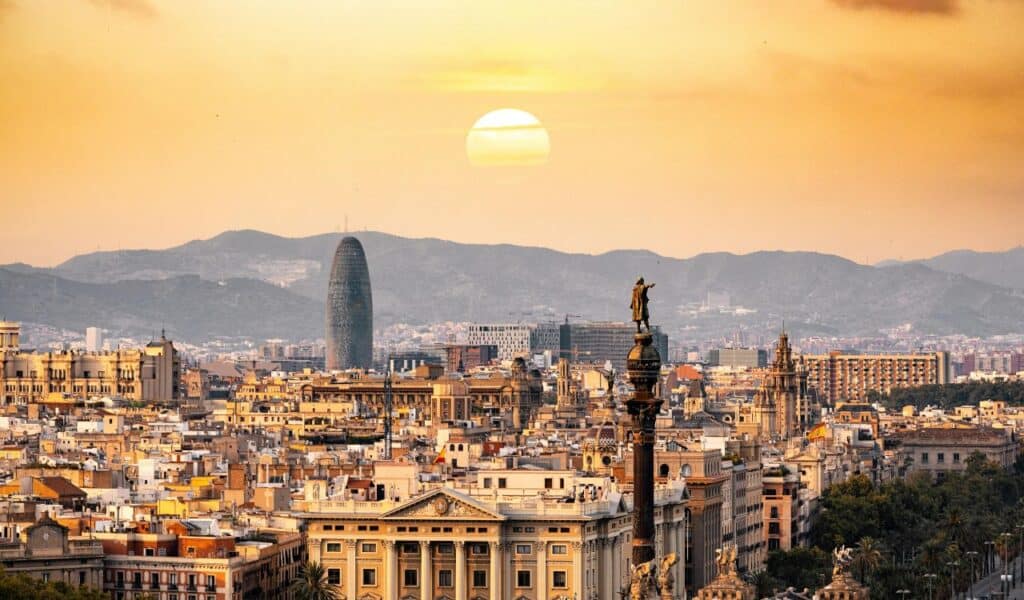 Panoramic view of Barcelona at sunset featuring historic buildings, the Columbus Monument, and mountains in the background under a golden sky.