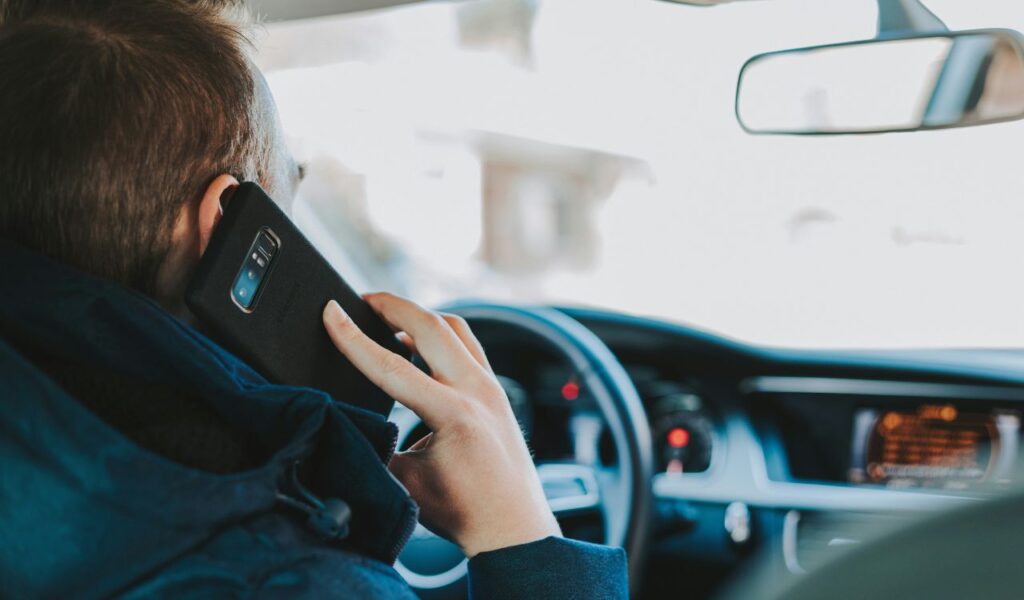 A man is seen from behind sitting in the driver’s seat of a car, holding a smartphone to his ear while driving, suggesting distracted driving behavior.