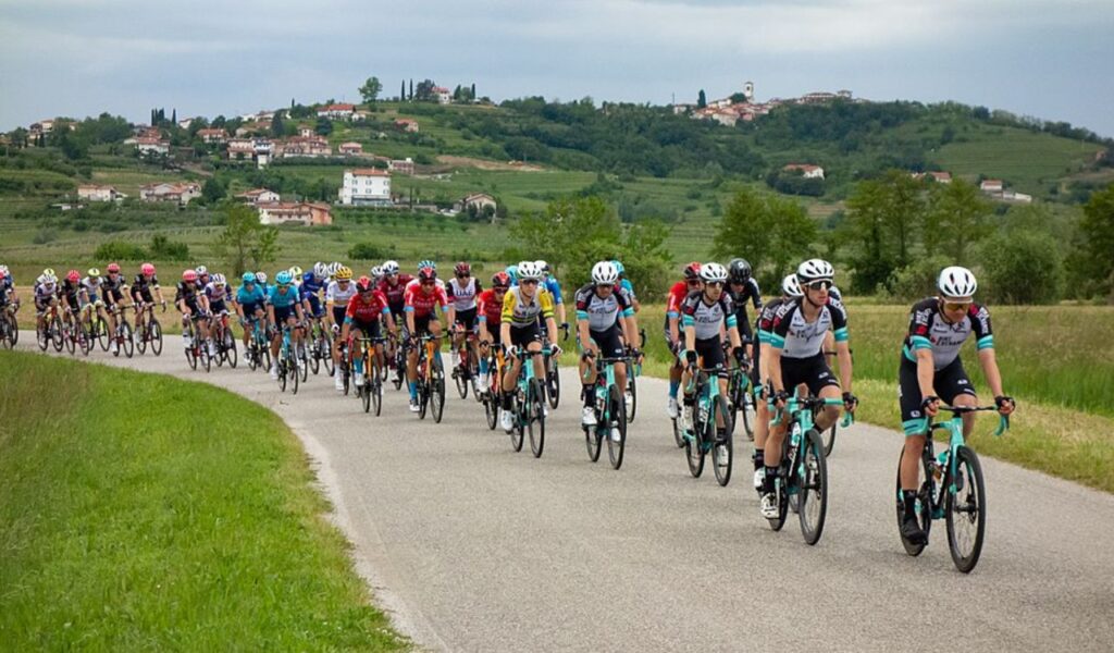 A large group of cyclists in professional gear rides along a winding rural road with green fields and hillside villages in the background.