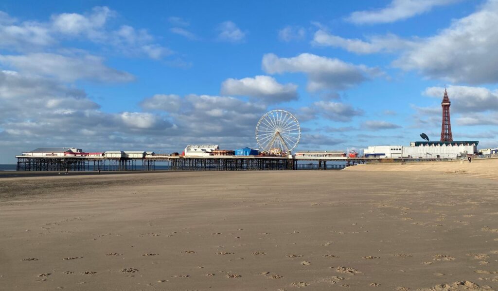A wide sandy beach leading to Blackpool Central Pier with a Ferris wheel, amusement park buildings, and the Blackpool Tower in the background.