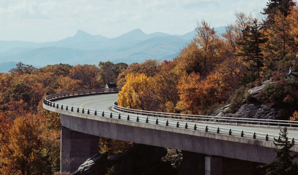 Blue Ridge Parkway curving through vibrant autumn foliage with distant mountain peaks in the background under a clear sky.