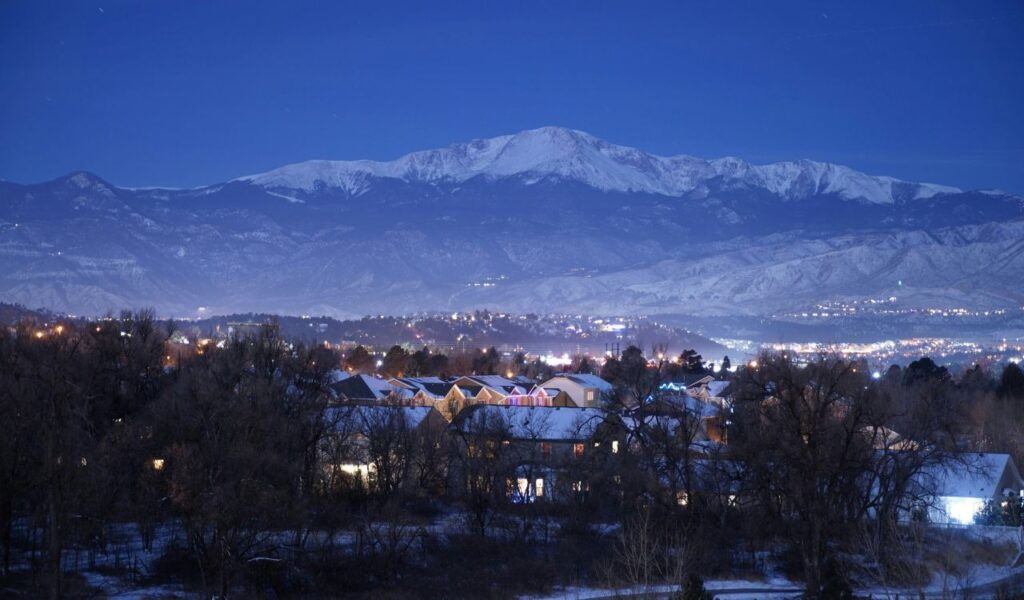 Snow-covered town at dusk with glowing houses in the foreground and a majestic mountain range in the background under a clear twilight sky.