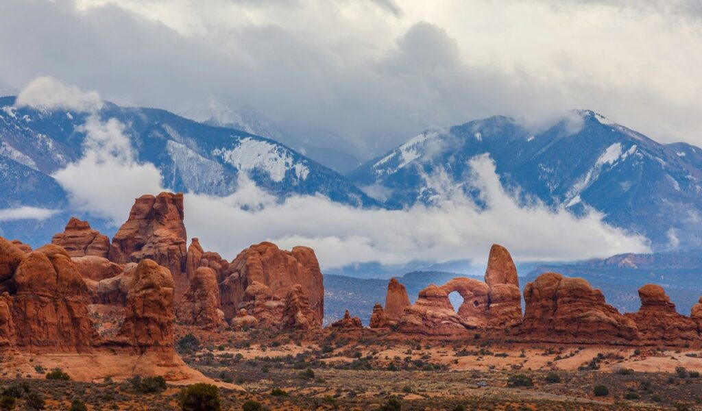 Red rock formations rise from the desert floor beneath dramatic clouds, with snow-capped mountains looming in the background.