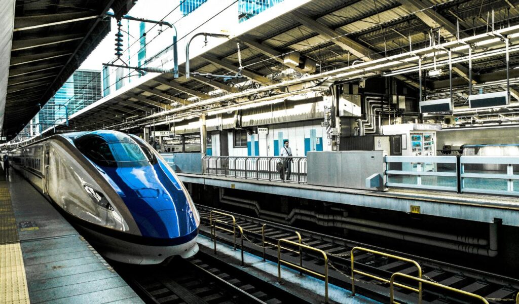 A sleek blue and silver Japanese bullet train stopped at a modern, high-tech train station platform with a few passengers waiting.