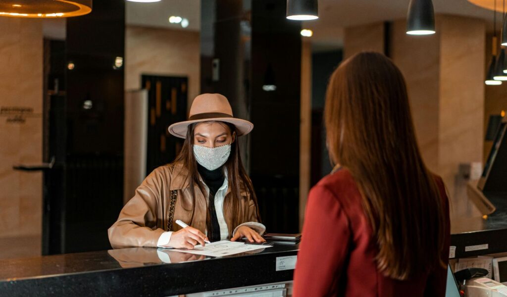 Woman wearing a mask and hat filling out paperwork at a hotel front desk while a receptionist assists her.