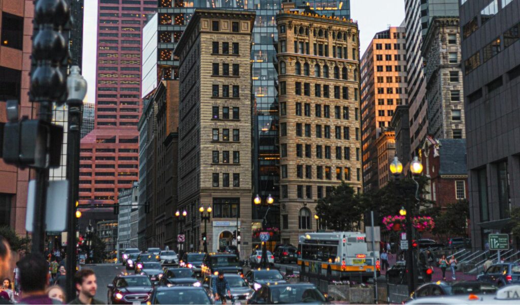 Busy downtown street in Boston, Massachusetts, with historic brick buildings, modern skyscrapers, traffic, and pedestrians during dusk.