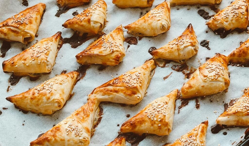 Golden-brown triangular bourekas topped with sesame seeds are arranged on a baking sheet lined with parchment paper.