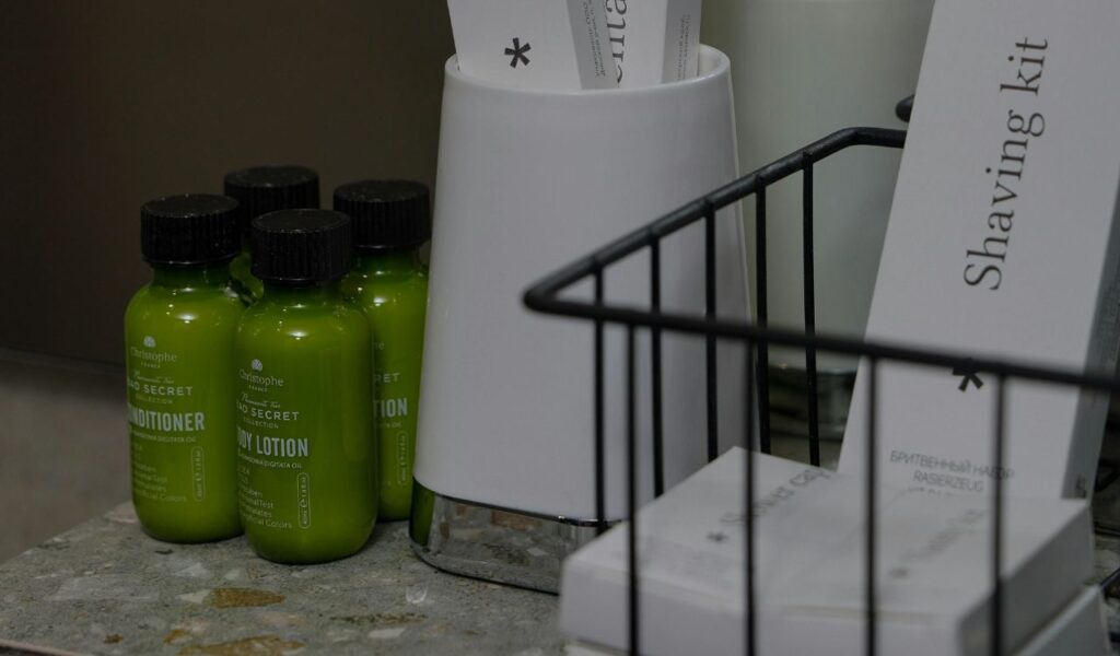 Close-up of hotel toiletries including green bottles of conditioner and lotion, a shaving kit, and grooming items on a bathroom counter.