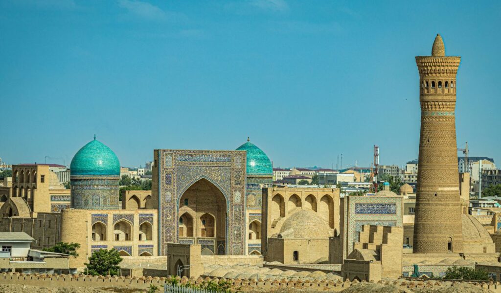 Historic Islamic architecture in Bukhara, Uzbekistan, featuring turquoise domes, intricate tilework, arched entrances, and the iconic Kalyan Minaret under a clear blue sky.