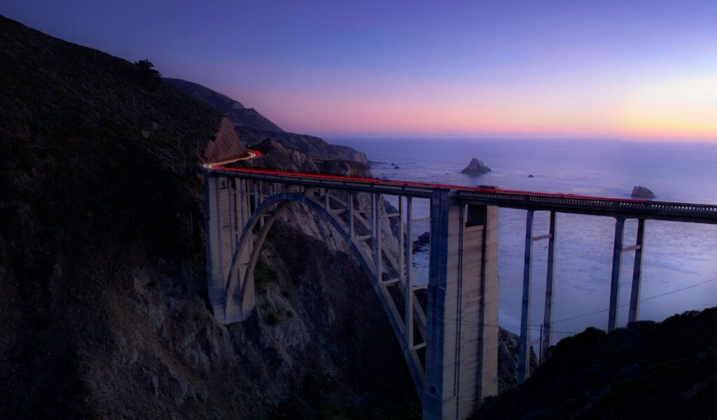 California Route 395 bridge at sunset, with car lights trailing across the span, rugged cliffs below, and the ocean stretching into the horizon.