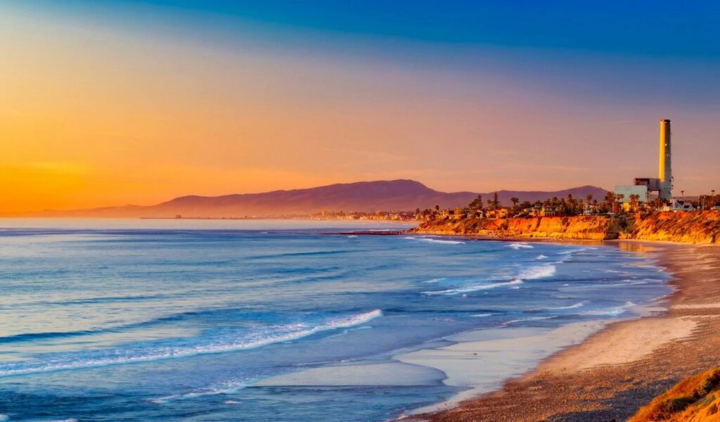 Golden sunset over Wright’s Beach in California with gentle waves, rugged cliffs, and a distant shoreline glowing in warm light.
