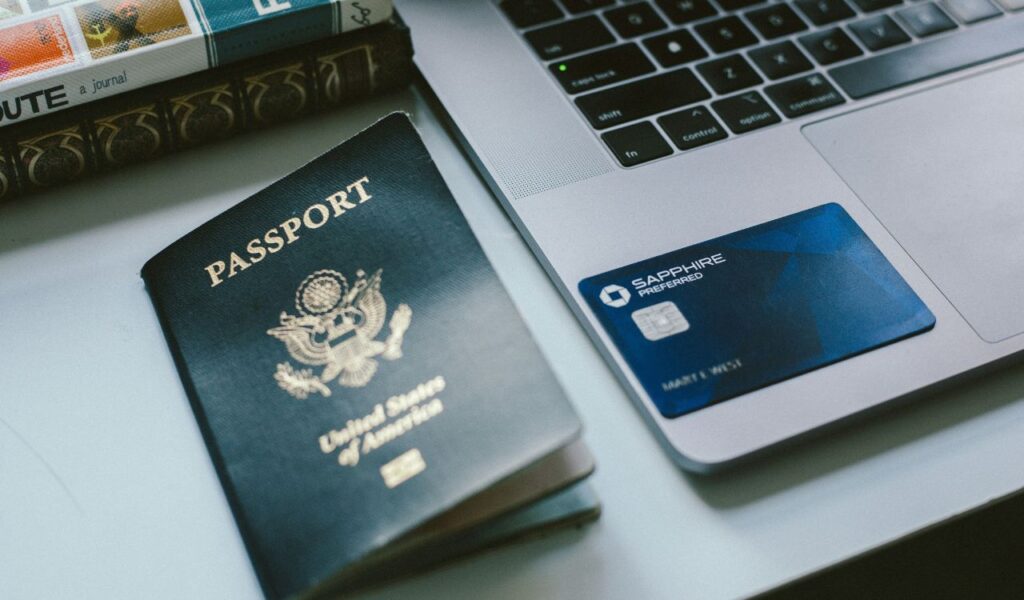 Close-up of a US passport, credit card, and laptop on a desk, suggesting important travel items kept together.