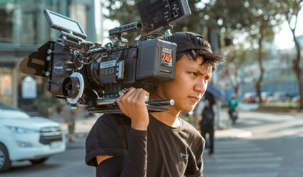 A young man in a black shirt carries a large professional film camera on his shoulder while walking through a city street.