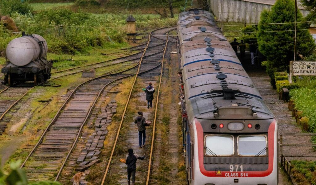 A modern passenger train at a rural station with people walking alongside the tracks, surrounded by greenery and old railcars.