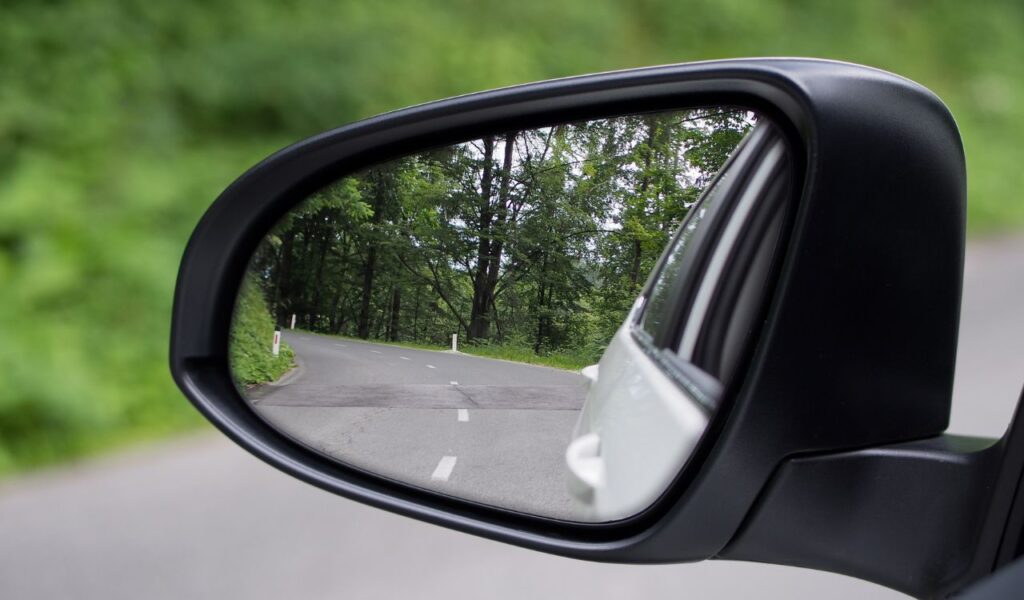 Curved road and trees reflected in a car's side mirror, emphasizing the importance of checking mirrors while driving.