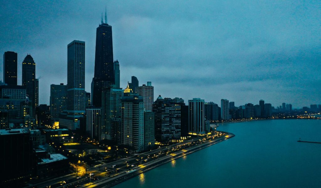 Aerial view of Chicago skyline at dusk with illuminated skyscrapers, Lake Michigan shoreline, and city lights reflecting off the water.