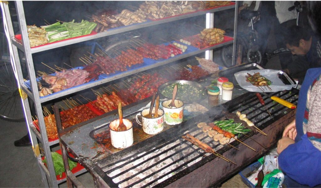 Chinese street food stall grilling skewers of meat and vegetables over charcoal in a bustling night market
