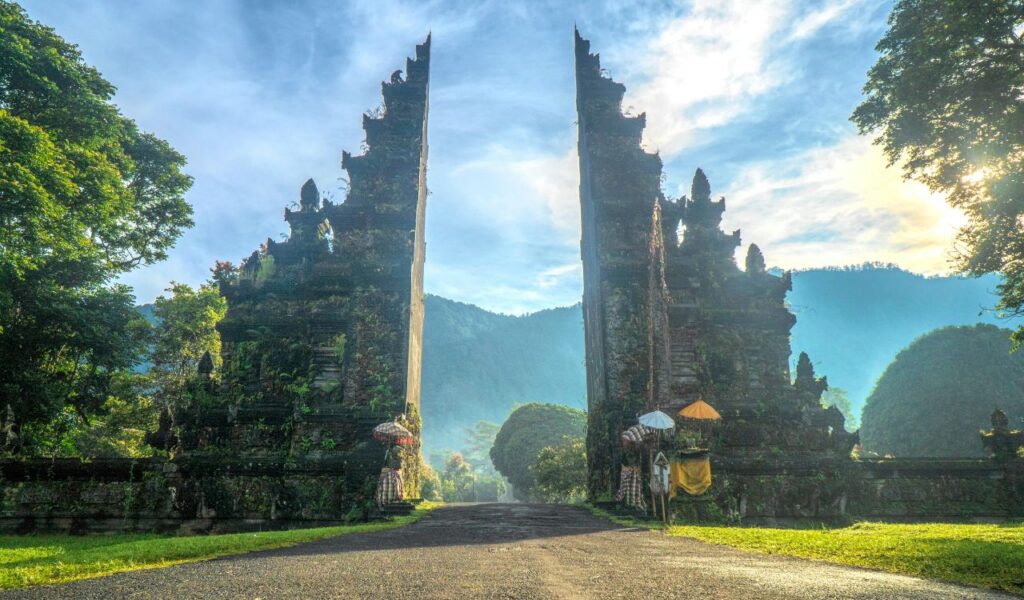 Balinese split gate with detailed carvings, framed by lush trees and misty mountains under a bright blue sky.