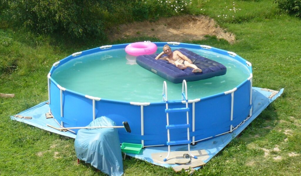 An older man lounges nude on an inflatable mattress in a small above-ground pool, surrounded by grass and trees.