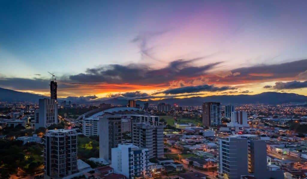 Vibrant sunset over San José, Costa Rica, with a glowing sky above the skyline, city lights beginning to twinkle, and mountains framing the horizon.