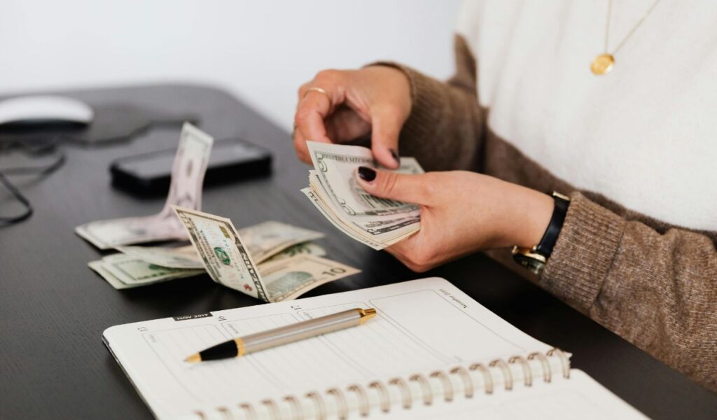Person counting U.S. dollar bills next to a planner, budgeting finances for travel or retirement expenses.