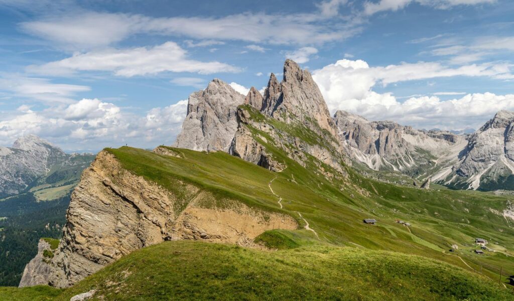 Hiking trail leading up to the jagged peaks of the Dolomites near Seceda, surrounded by lush green alpine meadows and scattered mountain huts under a bright blue sky.