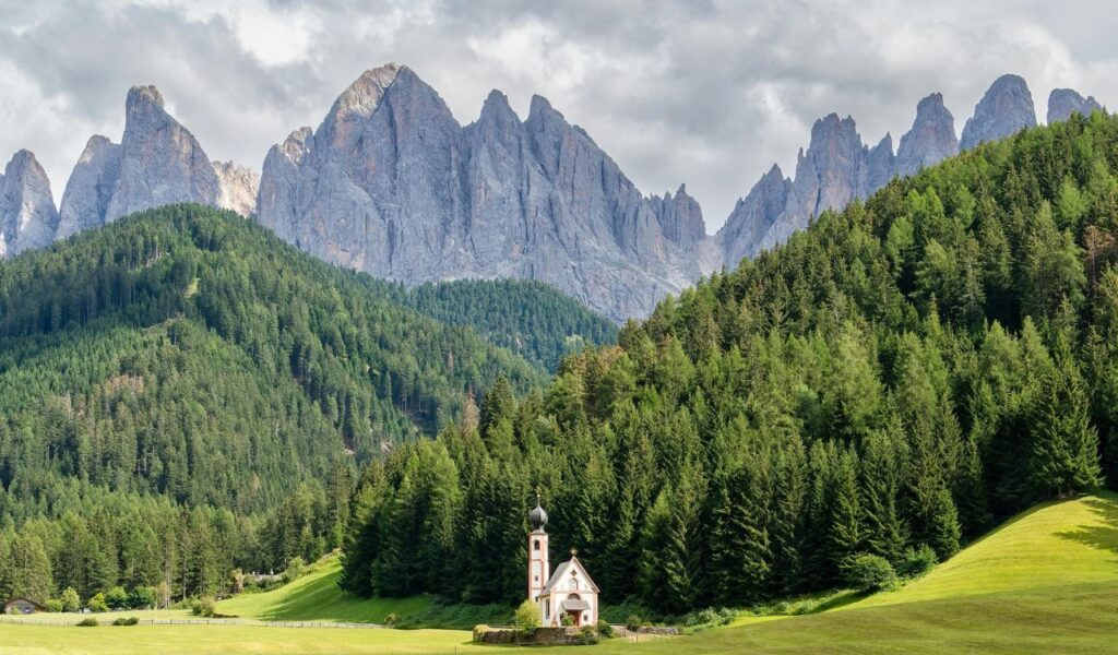 Small white church with a tall steeple sits in a green meadow, surrounded by dense pine forests, with the jagged peaks of the Dolomites rising dramatically in the background under a cloudy sky.