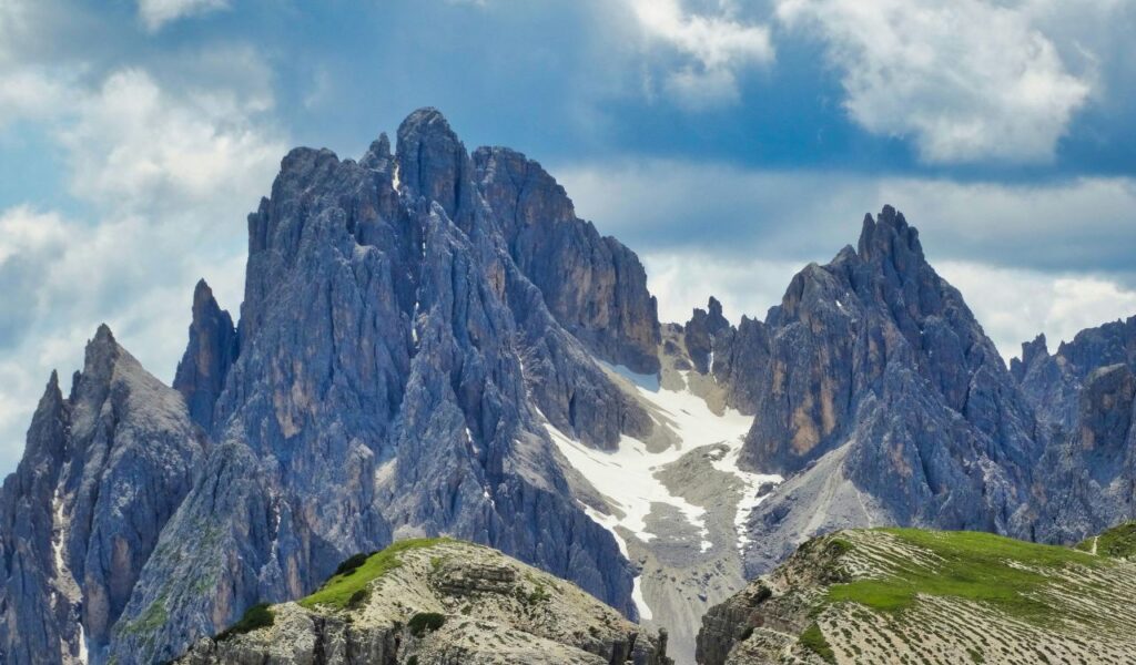 Jagged peaks of the Cadini di Misurina range in the Italian Dolomites, with streaks of lingering snow nestled between steep, rocky cliffs under a partly cloudy sky.