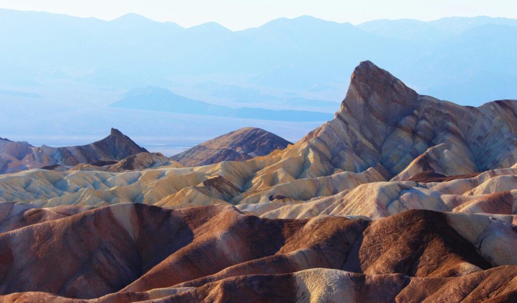 Colorful, jagged rock formations in Death Valley glow under soft sunlight with hazy mountain ranges in the distance.
