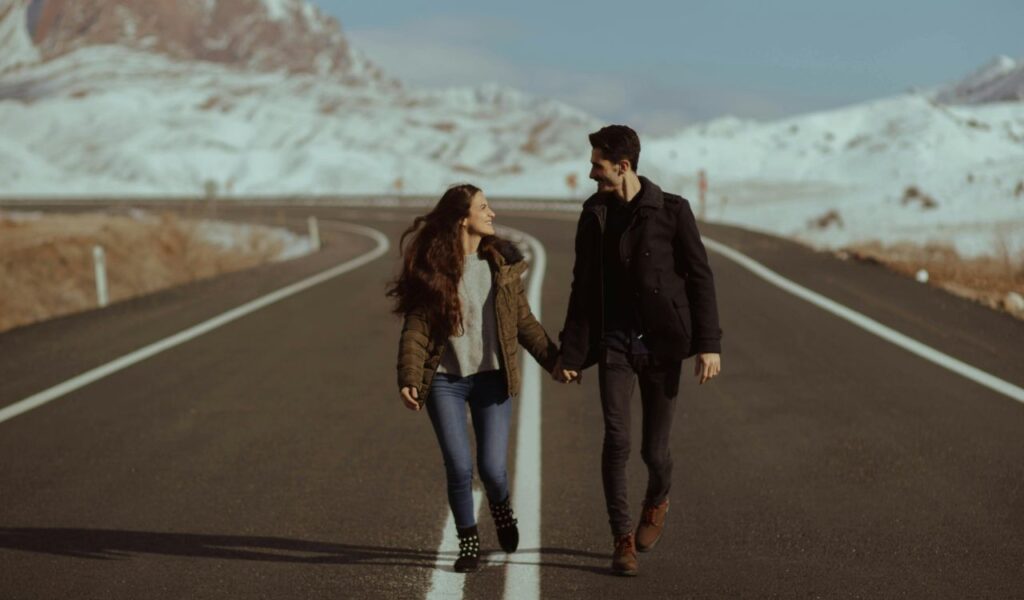 A couple walks hand in hand down an empty mountain road, smiling at each other with snow-covered peaks in the background.