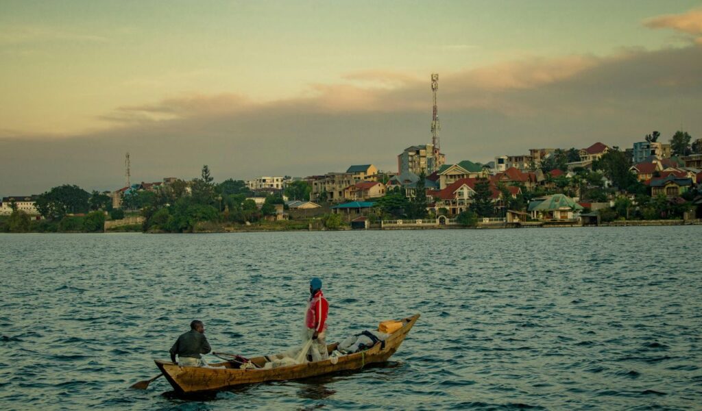 Two men in a wooden fishing boat on a lake with a colorful town and cell towers in the background at sunset.