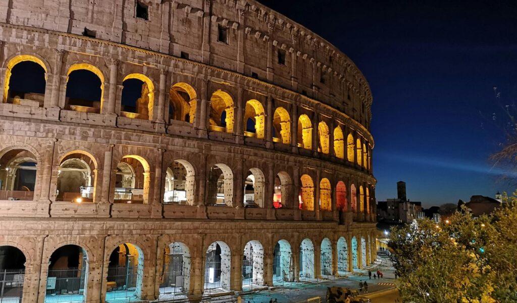 The Colosseum in Rome illuminated at night, with its iconic arches glowing in warm light against a dark blue sky and a few people nearby.