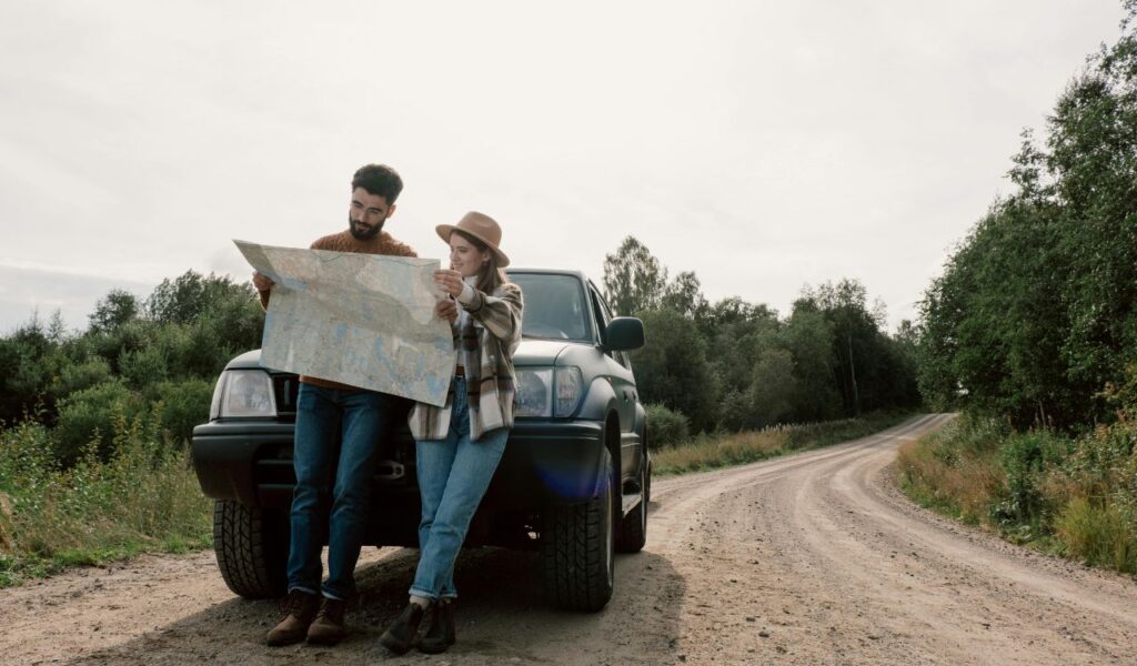 Couple standing by their SUV on a rural dirt road, reading a paper map, symbolizing road trip backup planning.
