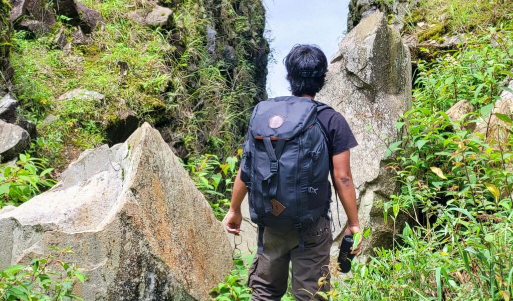 Solo backpacker standing between large rocks on a lush mountain trail, preparing to navigate an uncertain path.