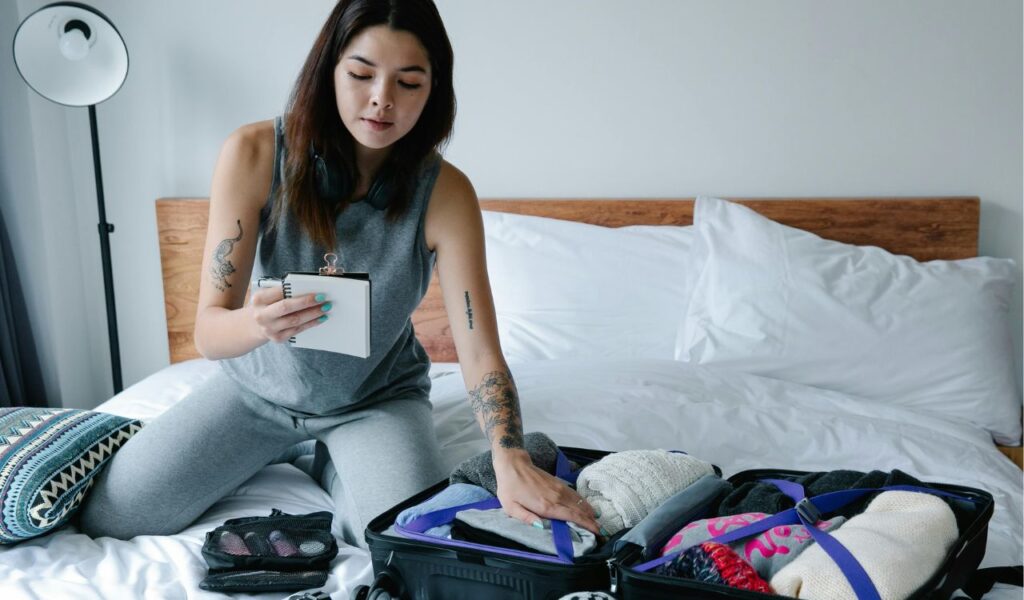 Young woman sitting on a bed, packing a suitcase and holding a notepad while organizing clothes and travel items.