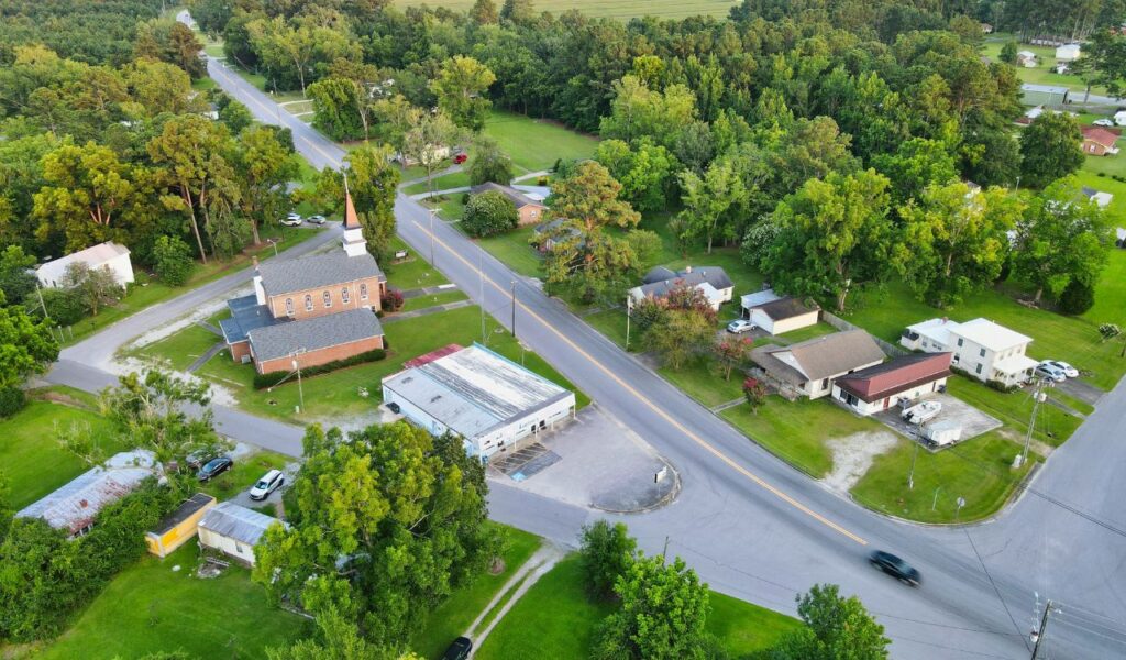 Aerial view of a small rural town with a church, houses, trees, and quiet intersecting roads surrounded by greenery and forest.