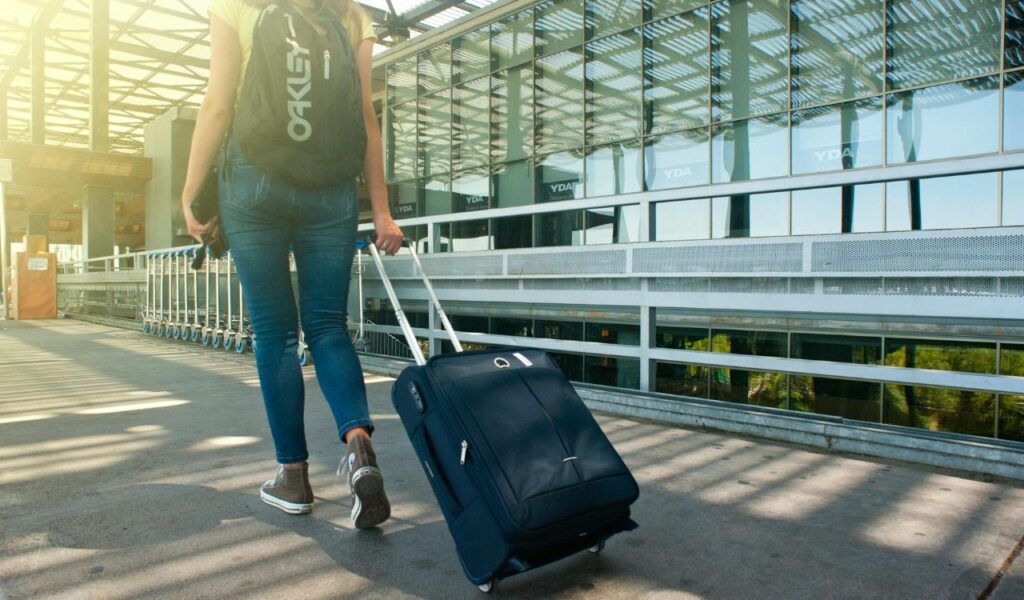 A traveler in jeans and sneakers pulling a large navy blue suitcase with one hand and wearing a black Oakley backpack at a bright, modern airport terminal.